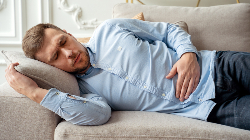 A young man lays on the couch at home holding his stomach in pain.