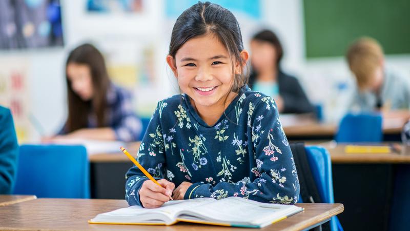 A young girl sits at a desk in a classroom at school and smiles for the camera.