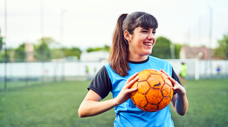 A young female athlete holds a soccer ball on an outdoor field.