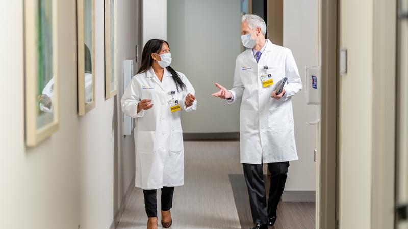 Two physicians, both wearing masks, walk down a hospital hallway as they talk.