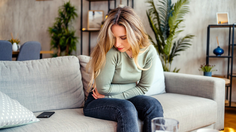 A young woman sits on her livingroom sofa and holds her stomach in pain.