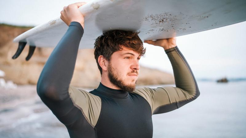 A young man holds a surf board over his head while standing on a beach.