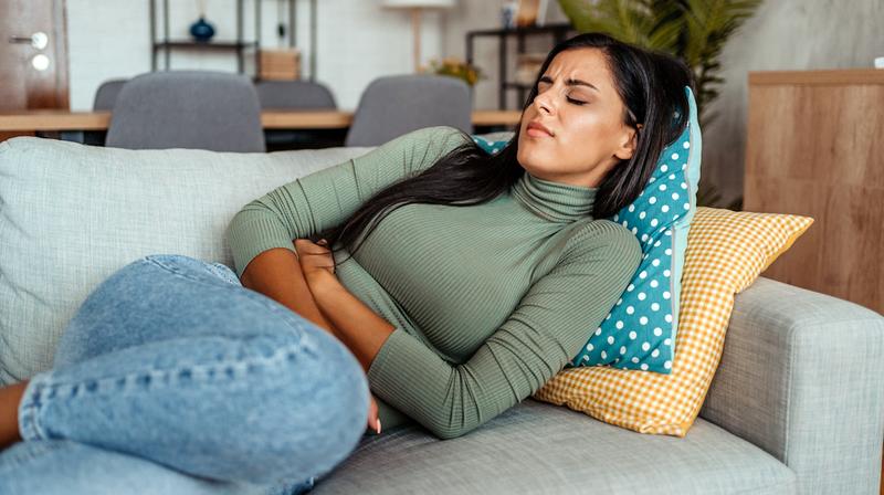 A young woman lays on the couch at home and clutches her abdomen in pain.