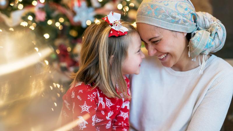 A woman wearing a head scarf touches foreheads with a young girl in front of a Christmas tree.