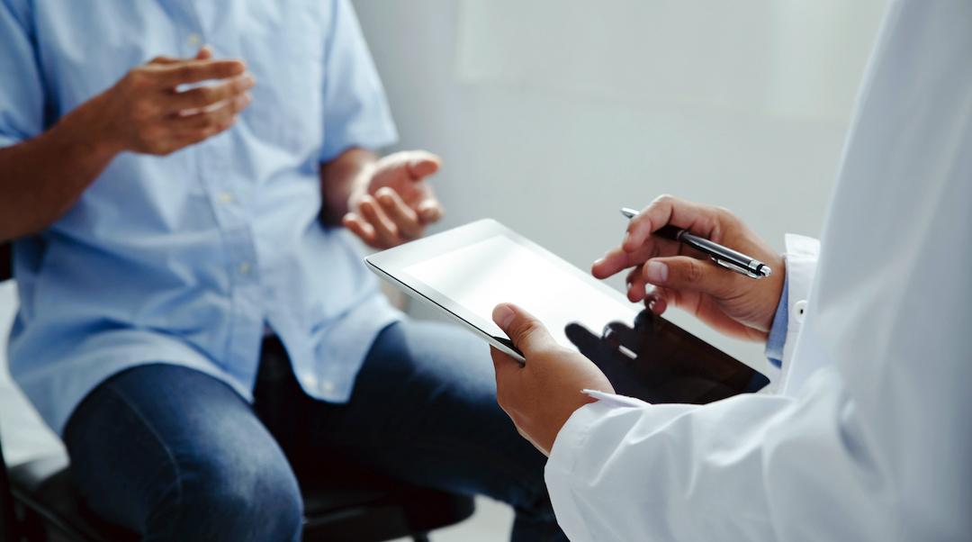 Close up of a doctor's hands holding an ipad with a patient sitting on an exam table in the background.