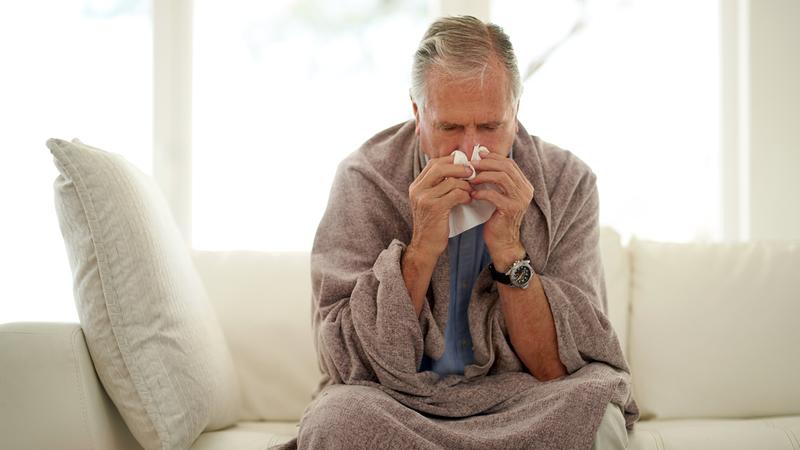 A senior man wrapped in a blanket blows his nose while recovering from the flu.