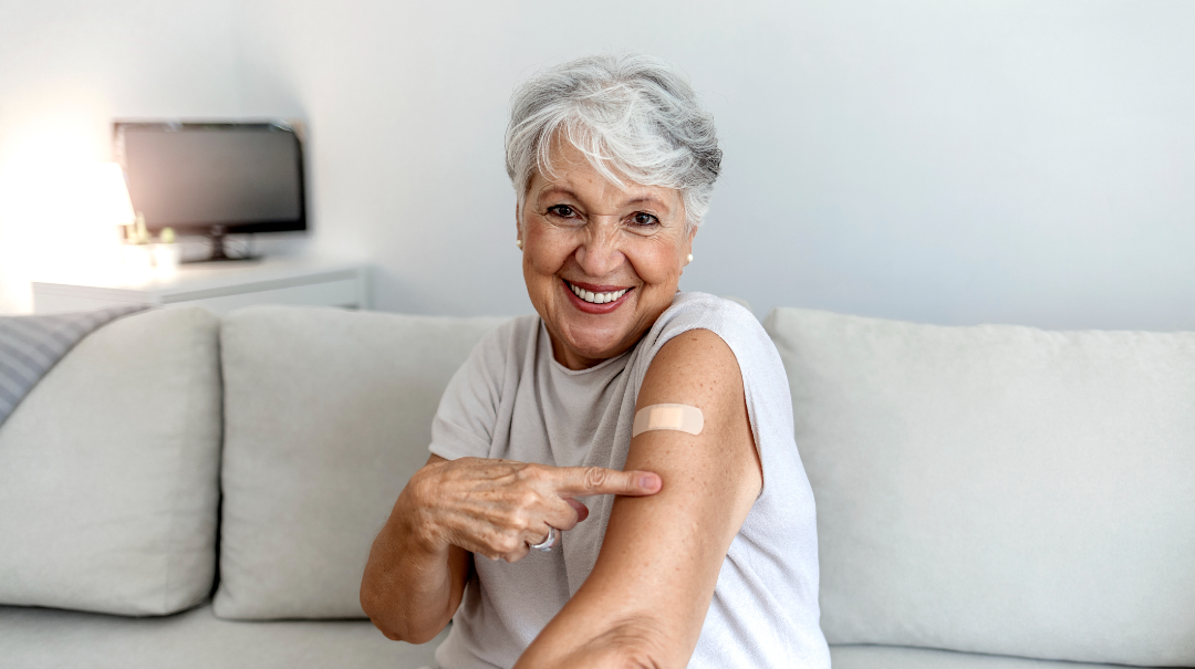 A senior woman sits on a sofa in her home and points to a band-aid on her arm indicating that she had been vaccinated.
