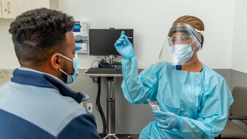 A healthcare provider wearing PPE swabs the nose of a male patient in a clinical setting.