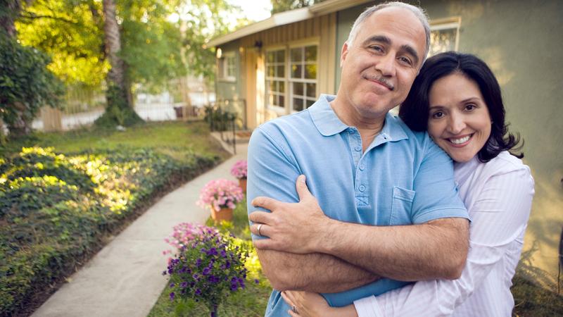 A wife hugs her husband while they both smile for the camera in front of their home.