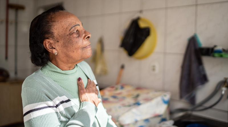 A senior woman stands in her kitchen with her hand on her heart.