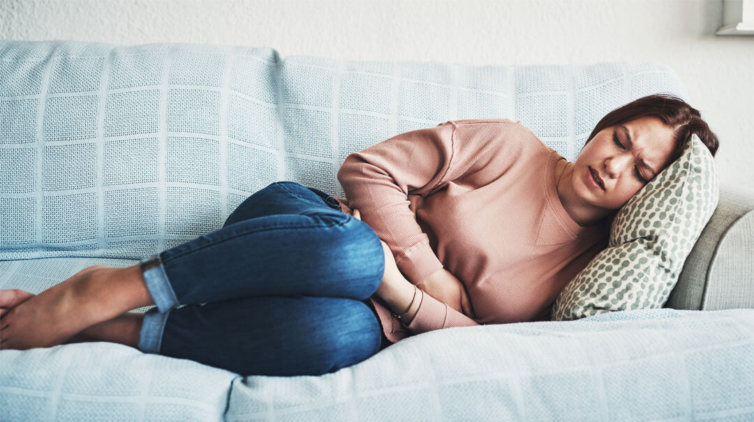 A woman lays on her couch at home and holds her stomach in pain.