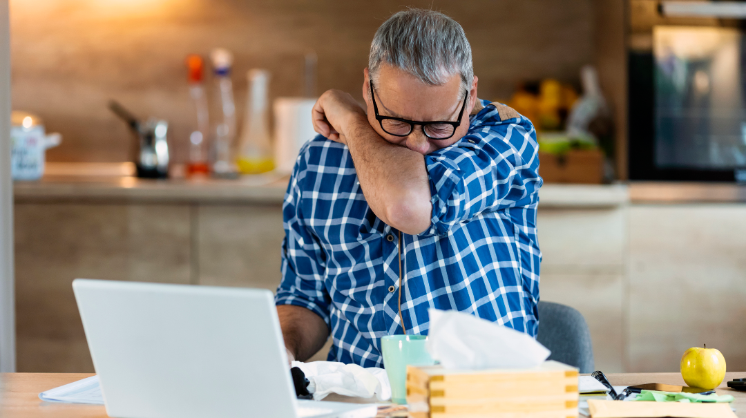 A man sneezes into his arm while working at his laptop at home.