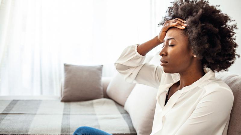 A woman sits on her living room sofa with her hand on her head and her eyes closed.