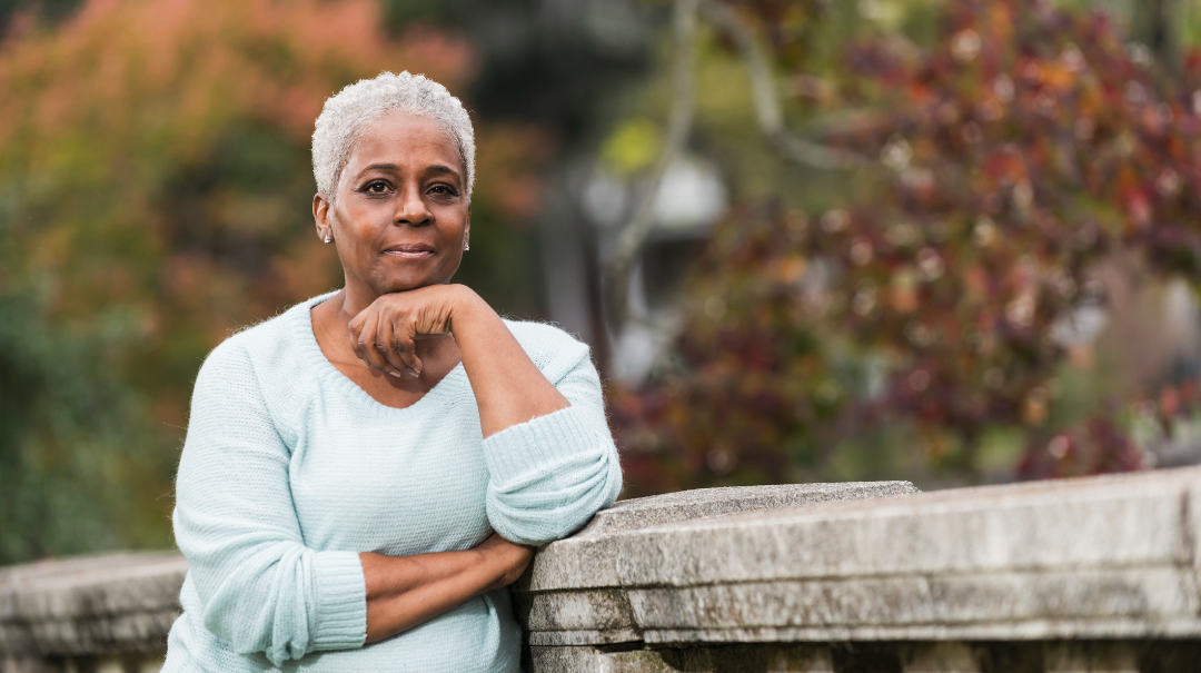 A woman leans on a wall in a garden and poses for a photo.