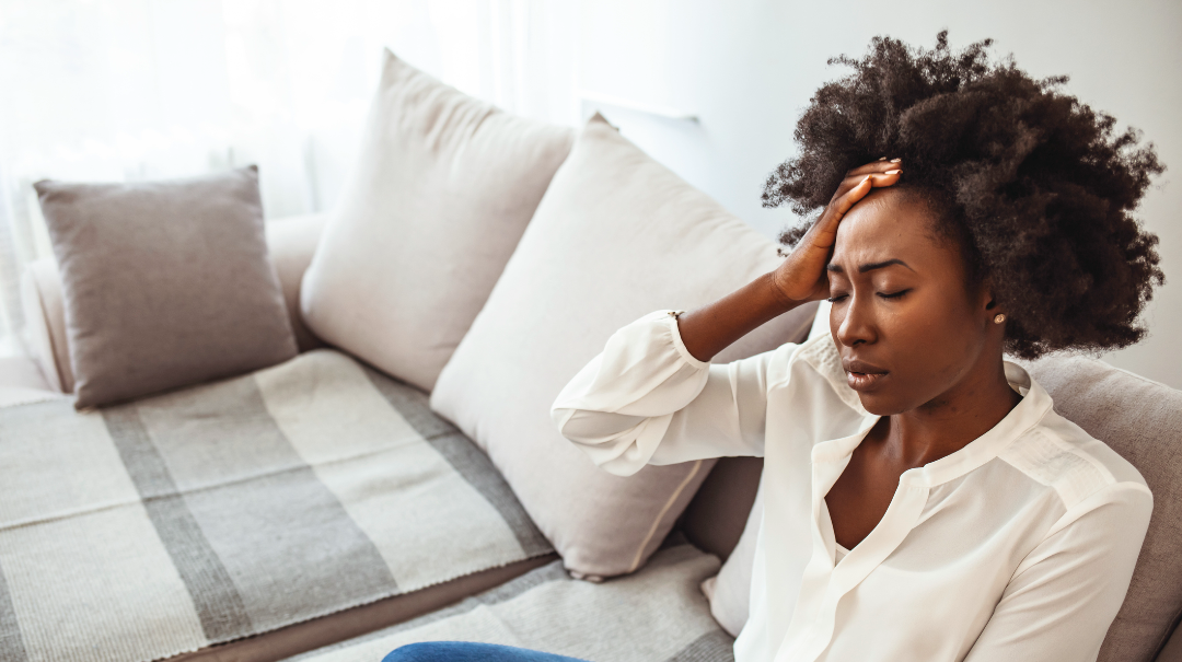 A woman sits on a couch in her home and winces in pain as she holds her hand to her head.