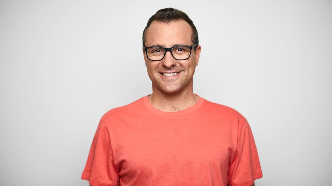 A middle-aged man in an orange shirt poses for a photo in front of a white wall.