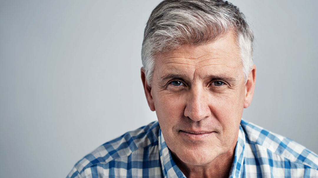 A mature adult man wearing a plaid shirt looks at the camera with a serious expression.