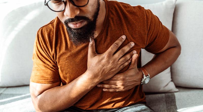 An African-American man clutches his chest while sitting on his livingroom sofa.