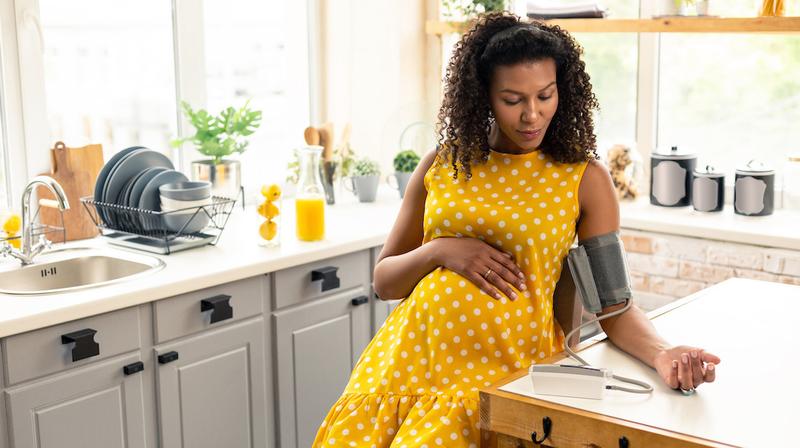 A pregnant woman takes her blood pressure reading while sitting in her kitchen.