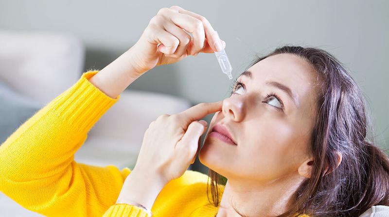 A woman wearing a yellow sweater puts drops in her eyes.