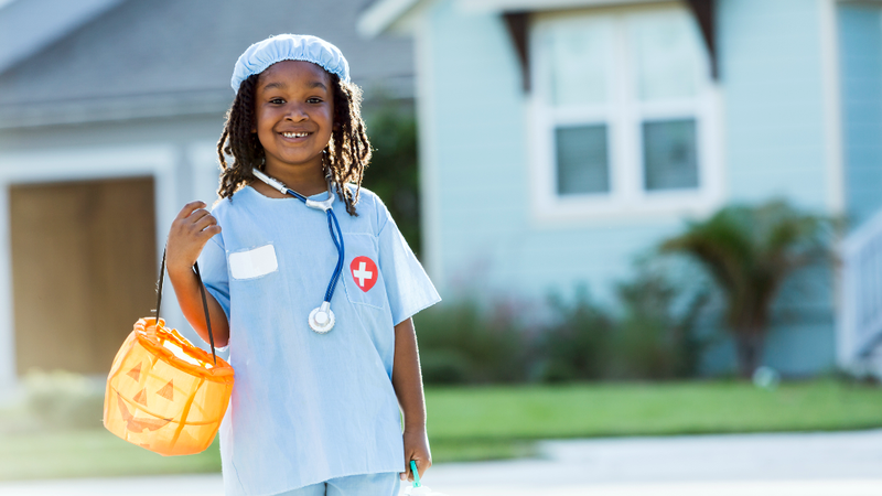 A young child dressed as a healthcare professional holds a halloween candy bucket while trick-or-treating.