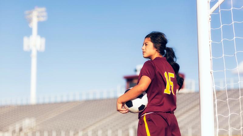 A female athlete holds a soccer ball on her hip in front of a soccer goal.