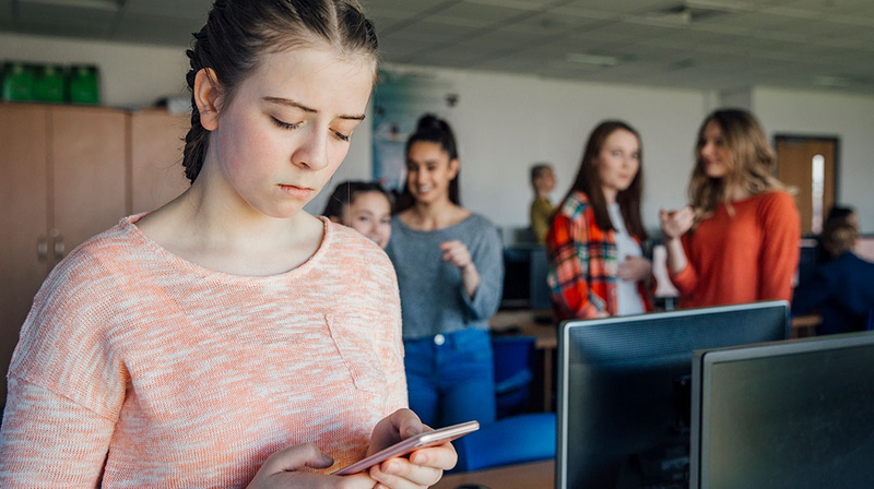 A teenaged girl texts on her phone at school, with kids in the background.