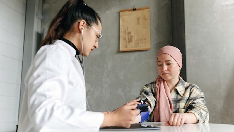 A doctor sits at a table with a young cancer patient and takes their blood pressure.