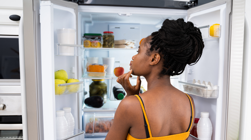 A woman stands in front of an open refrigerator to decide what to eat.