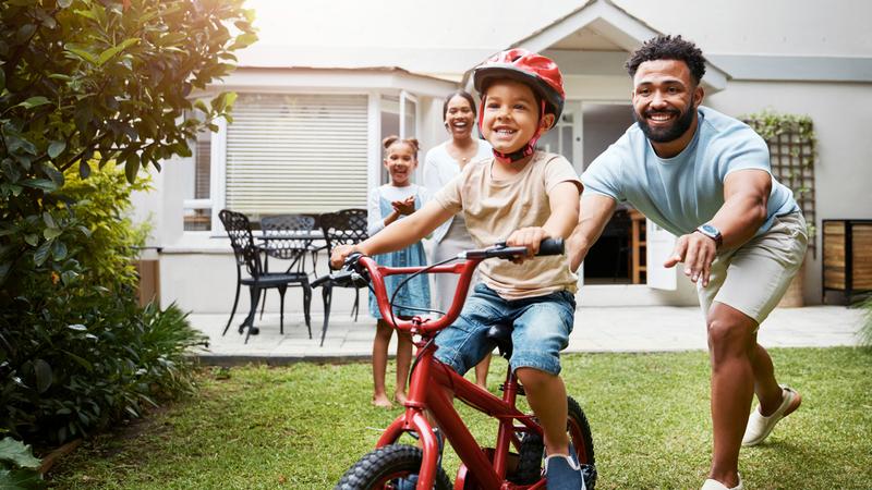 A father teaches his young son to ride a bike outdoors in front of their home.