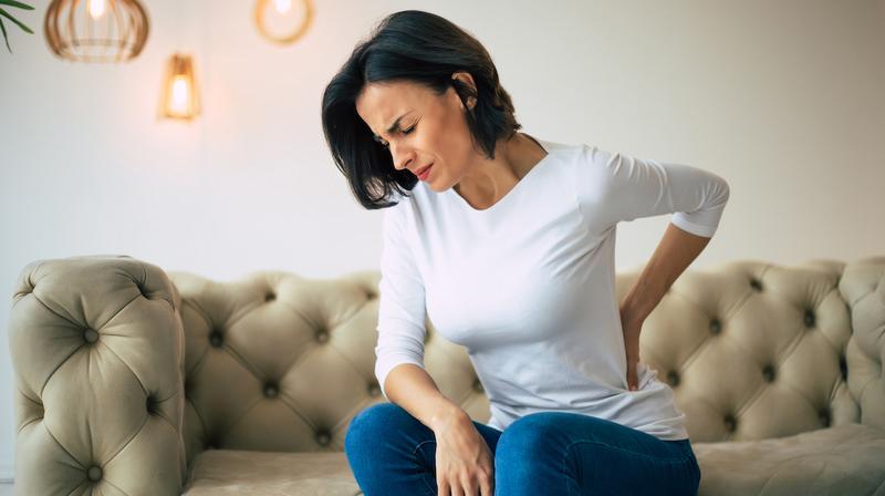 A woman sits on her livingroom sofa with her hand on her lower back wincing in pain.