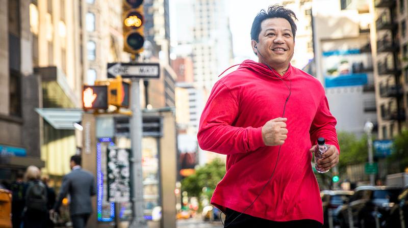 A man wearing a red jacket smiles as he jogs in a city setting.