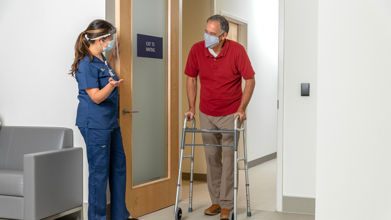 A healthcare worker, wearing a mask, holds the door open for a man using a walker.