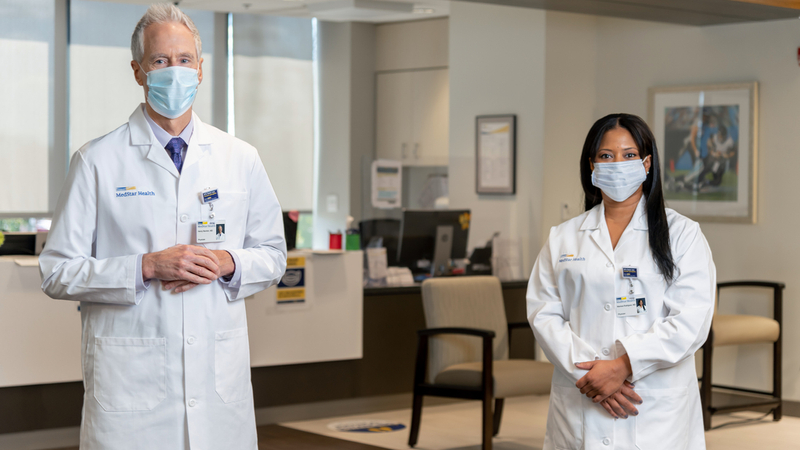 Two doctors, wearing masks, pose for a photo standing 6 feet apart.