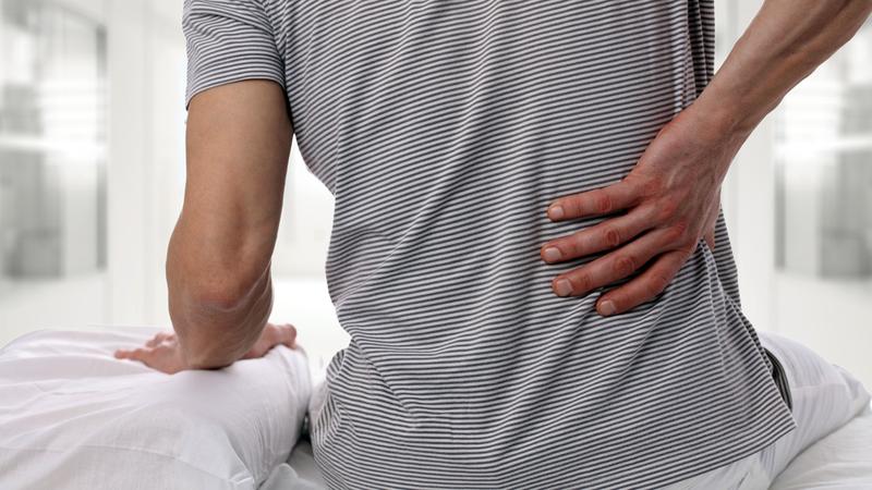 A man sits on his bed and puts his hand on his lower back indicating he is in pain.