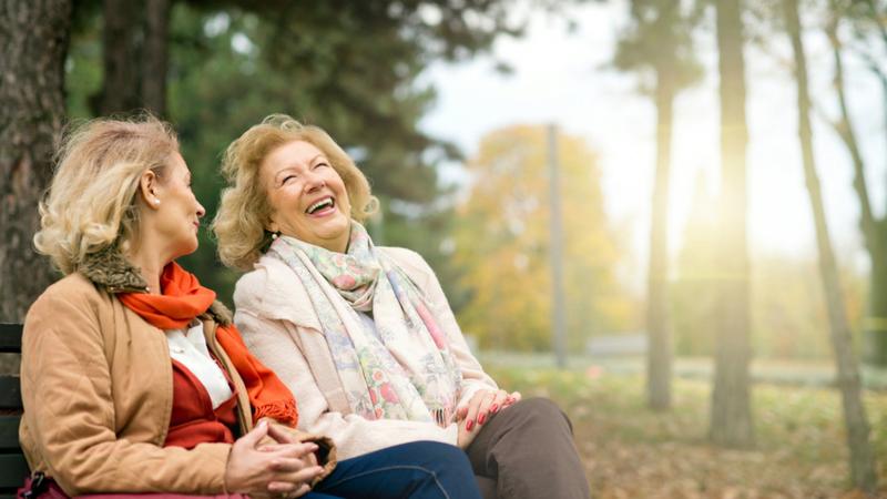 Two mature women laugh together as they sit on a park bench.