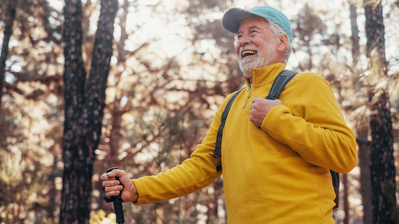 A senior man wearing a yellow sweater and a cap smiles while he walks through the woods.