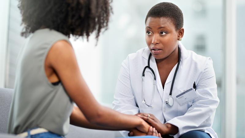 A doctor holds a woman's hand while talking with her in a clinical setting.