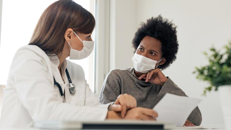 A doctor and patient talk in a clinical setting. Both people are wearing masks.