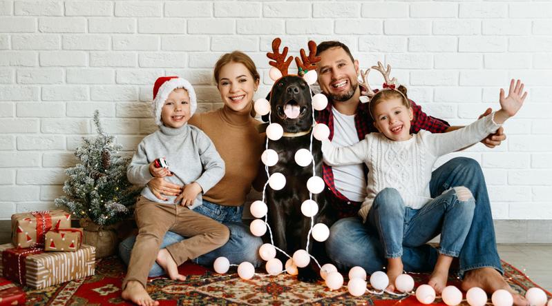 A family sits together on the floor and poses for a photo with their dog who is covered in Christmas decorations.