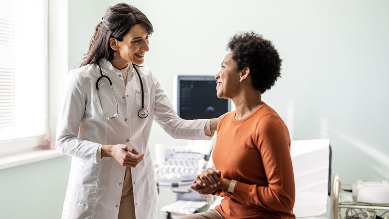 A doctor puts her hand on her patient's shoulder as they talk together in a clinical setting.