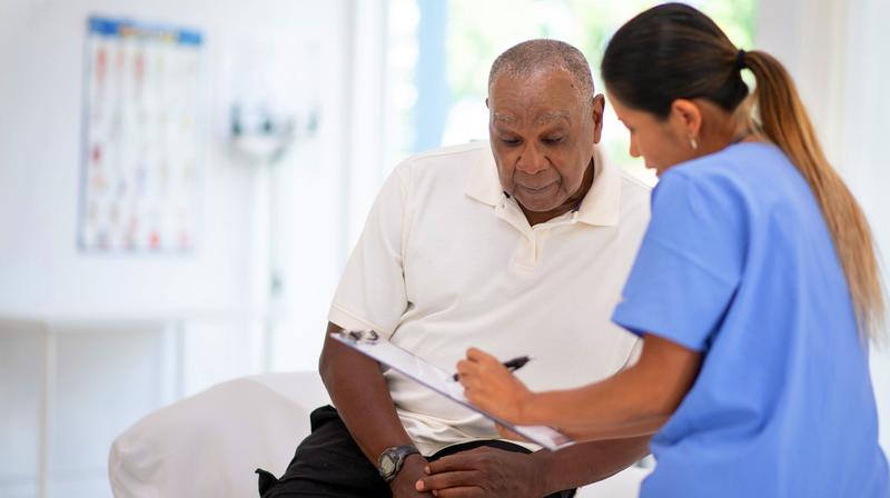 A man looks at papers on a clip board while a healthcare professional talks with him in a clinical setting.