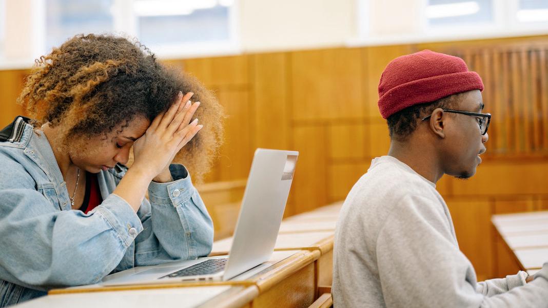 Two african-american students sit in a classroom - one has her head in her hands.