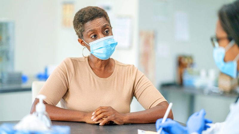 A senior woman wearing a mask, sits at a desk and talks with her doctor.
