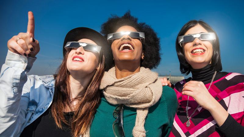3 women, wearing sun-safe glasses, look up at the sky during a solar eclipse.
