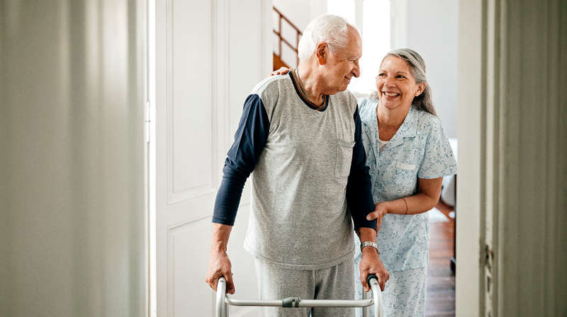 An elderly wife helps her husband walk with a walker at home.