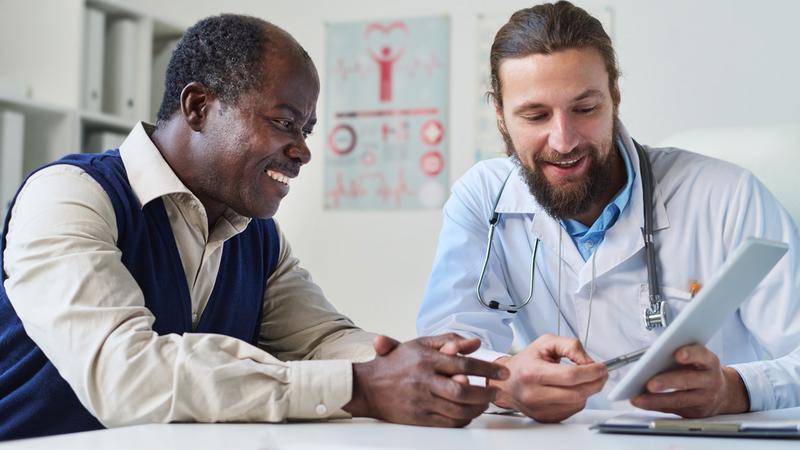 A doctor sits with a male patient and talks while showing him an ipad.