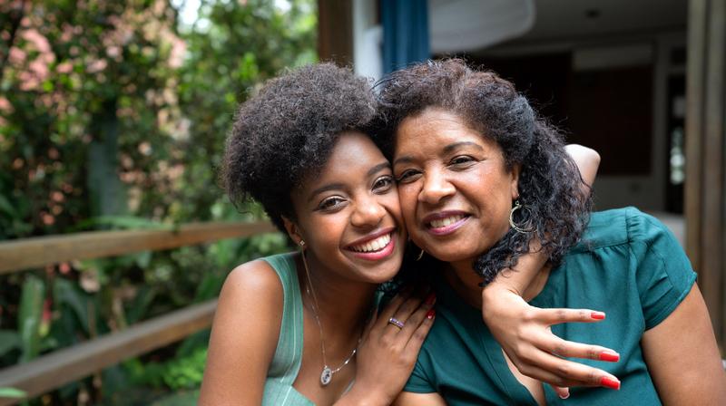 A daughter hugs her mother around the neck and they sit cheek to cheek as they smile for the camera outdoors. Both are wearing green shirts.