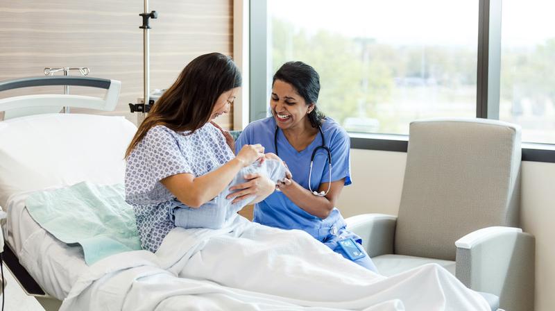 A nurse visits with a mother holding her newborn baby in a hospital room.
