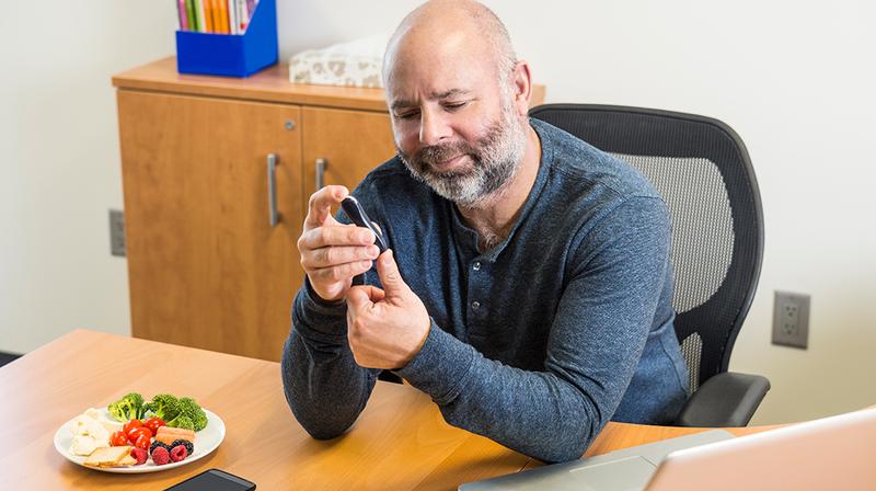 A man tests his blood sugar while sitting at a table preparing to eat a meal.